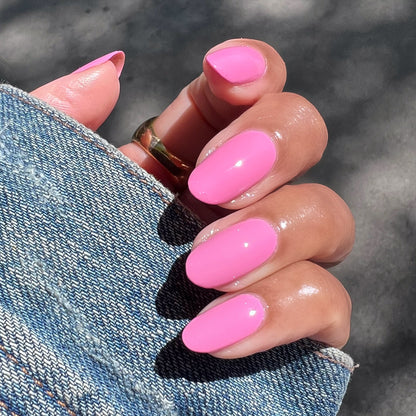 Hand with glossy bright pink Taffy gel polish on nails holding denim jacket sleeve in natural sunlight manicure view