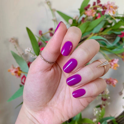Hand with nails painted vivid purple Grape Soda polish showing glossy at-home manicure with floral background