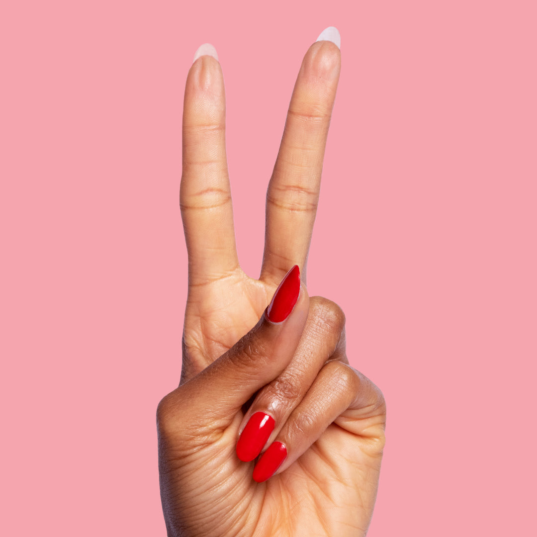 Hand showing peace sign with almond-shaped nails painted in glossy red Lollipop polish on pink background
